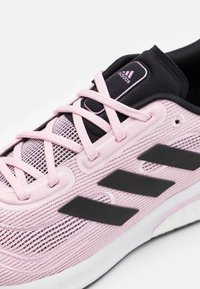 Zapatilla deportiva de color rosa claro con detalles en negro, parte superior de malla y cordones gruesos. Cuenta con tres franjas negras y suela acolchada.