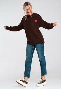 Brązowy sweter dzianinowy z różowym sercem, w połączeniu z niebieskimi dżinsami i wzorzystymi trampkami. Luźny krój i ściągacze.
