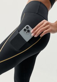 Crne atletske tajice s zlatnom prugom, s bočnom džepom u kojem se nalazi sivi pametni telefon. Materijal izgleda glatko i rastezljivo.