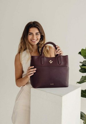 Dark burgundy leather handbag with dual handles, a front gold emblem, and a smooth texture, positioned on a white pedestal.