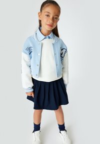 Giacca varsity azzurro chiaro con maniche bianche, dettagli ricamati, maglione bianco, gonna plissettata blu navy e sneakers bianche.