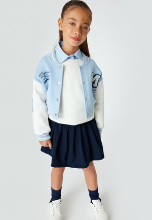 Giacca varsity azzurro chiaro con maniche bianche, dettagli ricamati, maglione bianco, gonna plissettata blu navy e sneakers bianche.