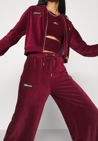 Survêtement en velours bordeaux avec veste cropped et pantalons à cordon. Comprend des patchs de marque en or, une texture côtelée et une coupe ajustée.