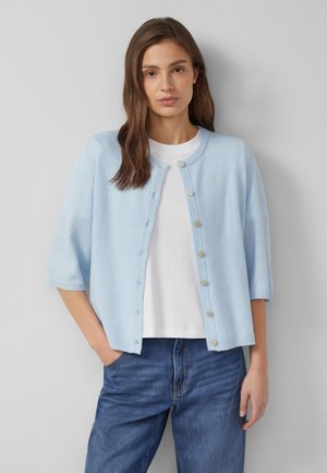 Junge Frau trägt eine hellblaue Strickjacke mit Knöpfen über einem weißen Hemd und blauen Jeans, steht mit einer Hand in der Tasche vor einer einfarbigen Wand.