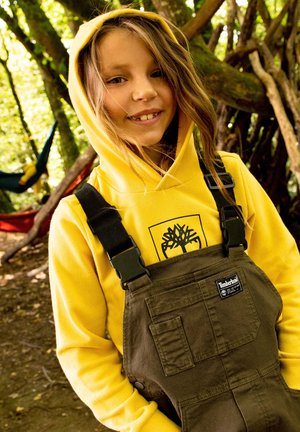 Un sweat à capuche jaune avec un logo, associé à des salopettes vert olive avec des poches et des bretelles réglables, dans un environnement naturel en plein air.