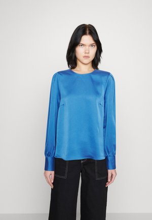 Blusa azul de satén de manga larga con escote redondo, textura suave y detalles de hombros fruncidos, combinada con pantalones anchos negros.