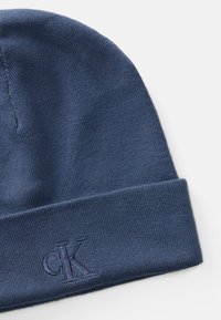 Marineblauwe gebreide beanie met opgevouwen rand, voorzien van een geborduurd "cK"-logo aan de voorkant tegen een witte achtergrond.