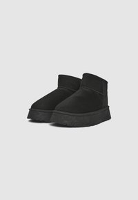 Bottines en suede noir avec un bout arrondi, des accents cousus et une semelle en caoutchouc épaisse et texturée. La semelle intérieure semble rembourrée pour plus de confort.