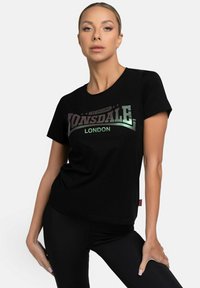 Juodas medvilnės marškinėlis, turintis atspindintį sidabrinį logotipą "Lonsdale London" per krūtinę, trumpomis rankovėmis ir laisvu siluetu.