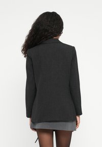 Kvinne med langt krøllete hår iført en mørk blazer og grå skjørt, vist fra ryggen mot en enkel hvit bakgrunn.