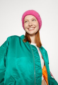 Turkos satäng bomberjacka med orange foder; rosa fluffig beanie; slät textur, dragkedja fram; figursydd design; har logotypdetalj på jackan.