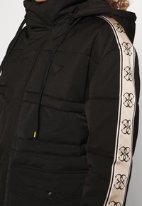 Giubbotto imbottito nero con colletto alto, caratterizzato da una striscia a pattern beige e nera lungo le maniche e con lacci regolabili sul cappuccio.