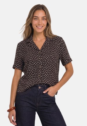 Femme souriante portant un chemisier noir à manches courtes avec des pois blancs, un jean foncé et un bracelet marron au poignet gauche.