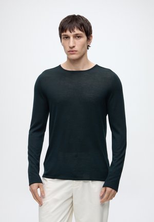 Man die een nauwsluitend zwart shirt met lange mouwen en witte broek draagt, staand tegen een effen lichte achtergrond, met het gezicht naar voren.