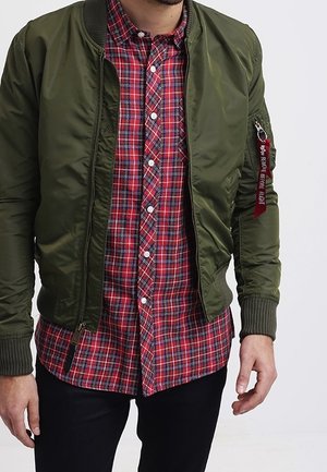 Mies, jolla on yllä avoinna oleva oliivinvihreä pommitakki, punainen ruudullinen napillinen paita ja mustat housut, seisoo yksinkertaisen taustan edessä.