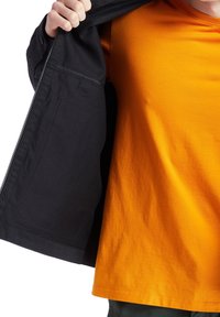 Černá bunda se zipovým uzávěrem a vnitřní kapsou, vrstvená přes jasně oranžové tričko. Důraz na texturu materiálu a kontrastní barvy.