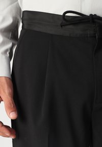 Zwarte broek van een gladde stof, met een brede tailleband met een satijnen afwerking en een trekkoordsluiting. Placketdetail zichtbaar aan de zijkant.