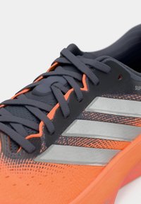 But sportowy z pomarańczową i ciemnoszarą cholewką o fakturze, z trzema srebrnymi paskami, zaokrąglonym kształtem i elastycznym materiałem.