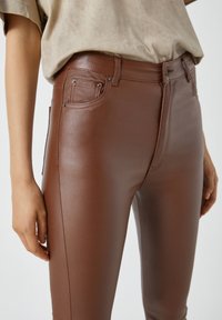Pantalon taille haute en simili cuir marron avec une texture lisse, un design à cinq poches et des accents en métal sur la taille et les poches.
