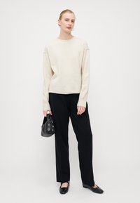 Maglione crema con scollo rotondo, vestibilità rilassata e orlo a costine, abbinato a pantaloni neri a gamba larga e una piccola borsa nera con dettagli ad occhio.
