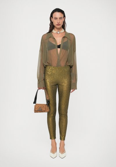 Femeie purtând o bluză transparentă măslinie peste sutien negru, pantaloni cu paiete aurii, tocuri albe, colier de perle, ținând o geantă cu imprimeu leopard pe fundal simplu.
