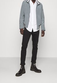 Man som bär grå jacka, vit skjorta med knappar, svarta byxor och svarta läderstövlar, står mot en helt vit vägg.