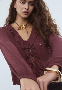 Blouse bordeaux avec des détails à volants et un décolleté en V, accompagnée d'un collier en cordon brun avec un accent de fleur jaune.