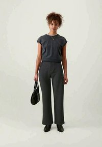 Donkergrijze top met korte mouwen, gecombineerd met hoog-gepolsterde, brede donkerkleurige broeken. Accessoires zijn een zwarte handtas en puntige schoenen.
