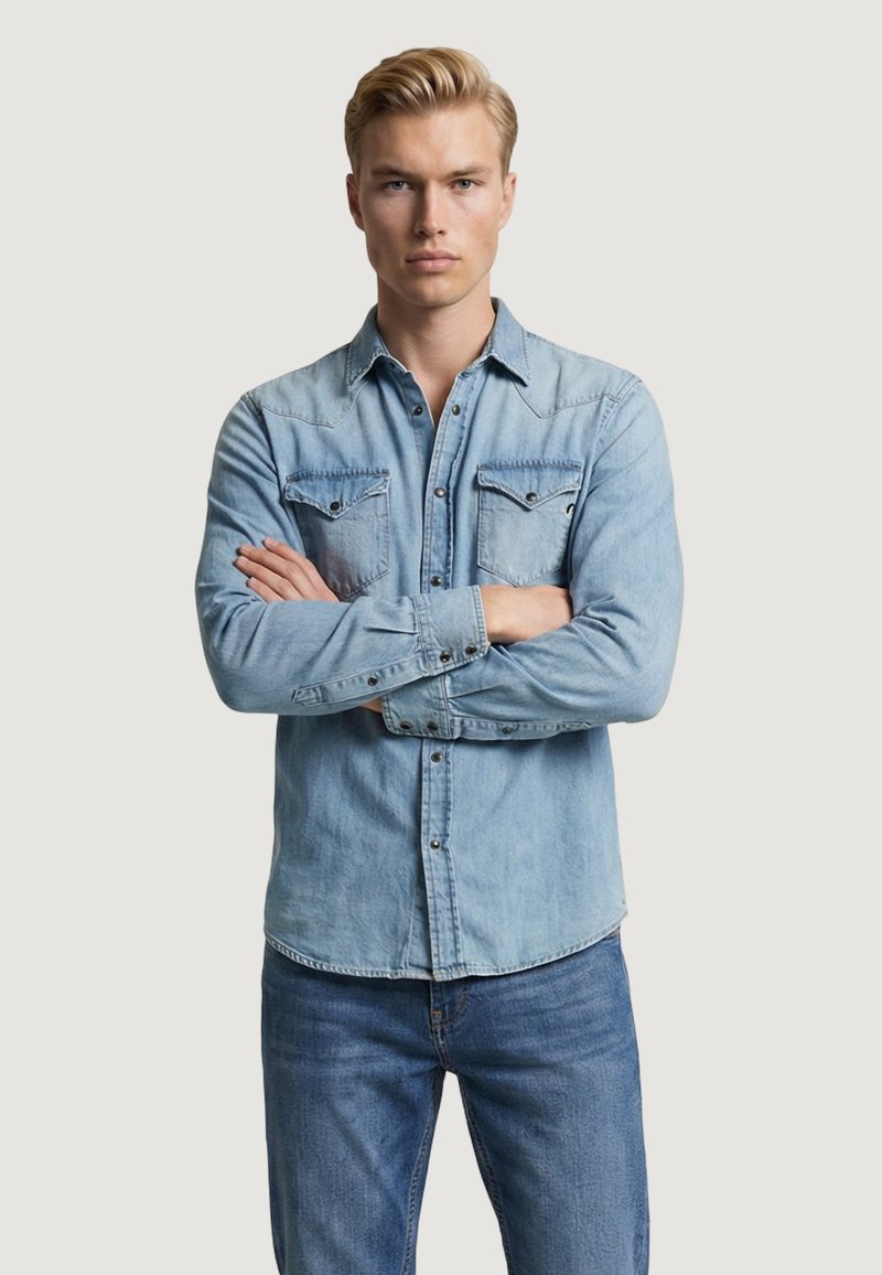 Joven con los brazos cruzados, vestido con una camisa de mezclilla azul claro y jeans, posando frente a un fondo claro y liso.