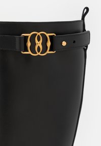 Ceinture en cuir noir avec une boucle double anneau dorée, présentant une texture lisse et une sangle réglable avec une fermeture à pression.