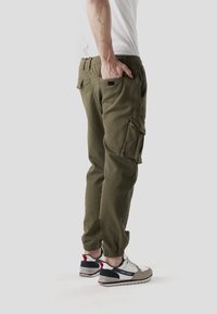 Pantalones cargo verde oliva con puños elásticos, múltiples bolsillos y un ajuste ligeramente holgado, combinados con zapatos deportivos gris claro con detalles en rojo.