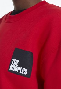 Sudadera roja hecha de tejido texturizado, con un parche cuadrado negro que lleva el texto blanco "THE KOOPLES" en el lado izquierdo.