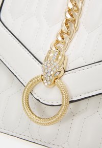 Valkoinen tikattu käsilaukku, jossa on kultaketju ja käärmeenkaltainen solki, jota koristavat strassit. Varustettu mustilla ompeleilla ja pyöreällä metallirenkaalla.