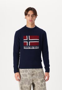 Marineblauer gestrickter Pullover mit einem Design der norwegischen Flagge in Rot, Weiß und Blau, mit gerippten Bündchen und Saum, kombiniert mit gemusterten beigen Hosen.