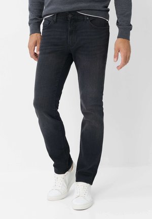 Mann trägt dunkle Slim-Fit-Jeans, weiße Turnschuhe und ein dunkelgraues Langarmshirt und steht vor einem hellen Hintergrund.