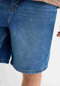 Shorts de mezclilla en azul claro con un corte clásico, que cuentan con un dobladillo sin terminar y costuras visibles. La textura de la tela es resistente y ligeramente desgastada.
