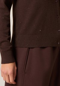 Cárdigan de punto marrón con dobladillo y puños acanalados, que presenta pequeñas motas de textura y cierre de botones. Combinado con pantalones marrones oscuros.