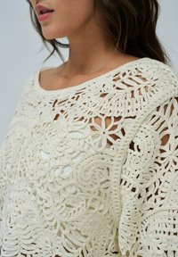 Cremfarbener Häkeloberteil mit filigranen Blumen- und Wirbelmustern, weitem Ausschnitt und lockerer Passform. Strukturierter Stoff mit offenem Gewebedesign.