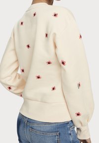 Sweat-shirt crème avec broderie d'araignée rouge et noire, col rond et poignets côtelés. Coupe décontractée avec un tissu texturé.