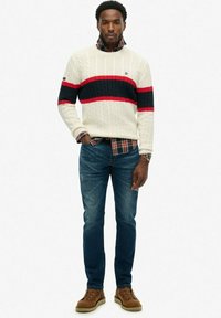 Pull en maille torsadée crème avec des rayures bleu marine et rouge, superposé à une chemise à carreaux. Porté avec un jean bleu et des bottes marron, se tenant sur un fond neutre.