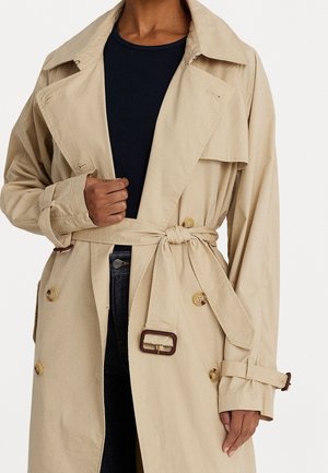 Persona que lleva un abrigo beige con cinturón anudado sobre una camiseta negra y pantalones oscuros, sujetando la solapa del abrigo con una mano.
