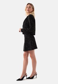 Zwarte tweed suit met een kort jasje en een uitlopende rok, met metalen draadaccenten. Model draagt zwarte slingback hakken.