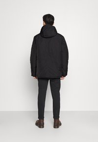 Černá vyp padded bunda s vysokým límcem, nastavitelnou kapucí a bočními kapsami, kombinovaná s tmavými kalhotami a hnědými botami. Jednoduchý design.