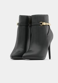 Botines de tacón alto de cuero negro con correas de hebilla dorada y punta afilada sobre un fondo blanco.