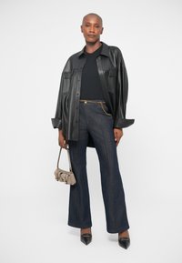 Veste chemise en cuir noir, haut noir, pantalon flare en jean noir Taille haute avec détail en chaîne, et escarpins noirs à bout pointu. Petit sac à main beige.