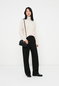 Kvinne som har på seg en kremfarget genser med høy hals, svarte brede bukser, svarte loafers, og bærer en liten svart crossover-veske på en hvit bakgrunn.