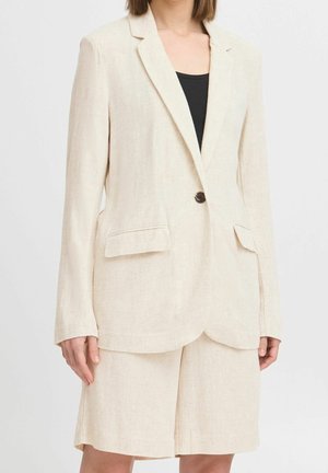 Kobieta ubrana w kremową lnianą marynarkę z jednym guzikiem i pasujące krótkie spodenki do kolan na czarnej bluzce, stojąca na jednolitym tle.