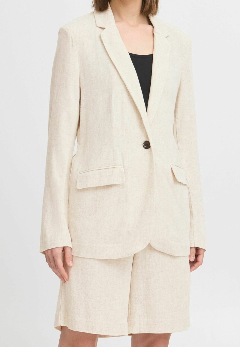 Femme portant un blazer en lin crème avec un bouton et un short assorti mi-cuisse sur un haut noir, debout devant un fond uni.