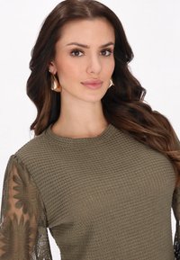 Pulover verde măsliniu cu corp tricotat texturat și mâneci din dantelă florală transparentă. Modelul poartă cercei îndrăzneți, reflectorizanți.