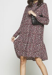 Květinové šaty s vysokým límcem, dlouhými rukávy a sukni s volány v červené, bílé a modré barvě. Doplnené malou černou texturovanou kabelkou.